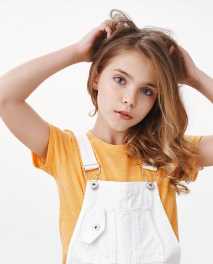 Tender lovely young blond girl in summer dungarees over yellow t-shirt, look thoughtful and cute camera, scratch hair bothered, touch hairstyle, suffer head lice, lacking vitamins, white background.